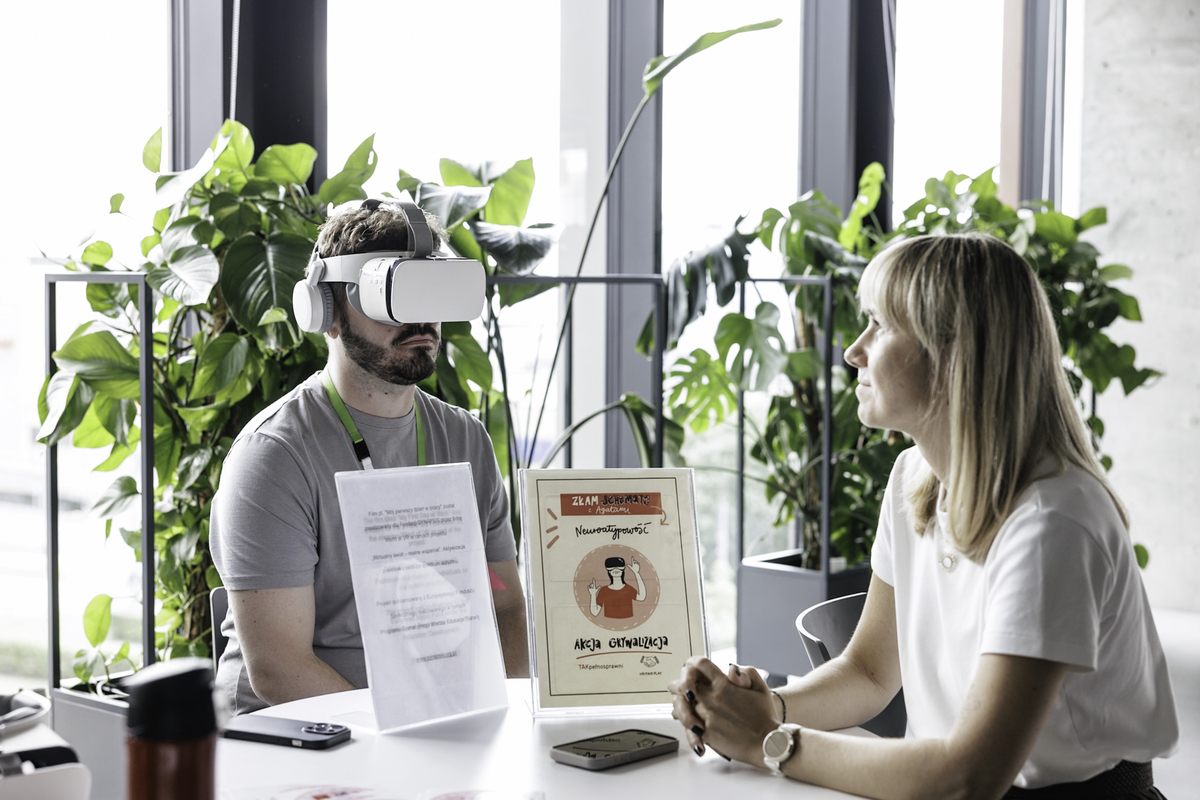 A man wearing VR goggles sits at a table with a sign reading “Activation Action - Neuroatypism.” On the other side, a woman from the Foundation's team is talking to a participant.